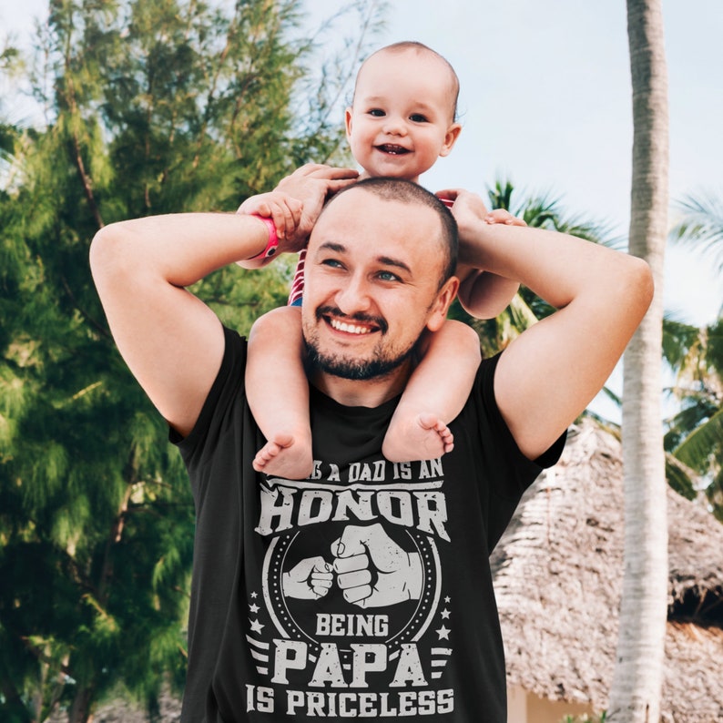 K&ouml;nnte beinhalten: Ein schwarzes T-Shirt mit einem wei&szlig;en Grafikdesign, auf dem "Ein Vater zu sein ist eine Ehre, Papa zu sein ist unbezahlbar" steht. Die Grafik zeigt auch zwei sich sto&szlig;ende F&auml;uste. Ein Mann tr&auml;gt das Shirt und tr&auml;gt ein kleines Kind auf seinen Schultern.