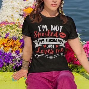 Puede incluir: Camiseta negra con el texto "I'M NOT SPOILED MY HUSBAND JUST Loves me" en rojo y blanco. El dise&ntilde;o incluye un beso de l&aacute;piz labial rojo y corazones. La persona lleva pantalones rosas y est&aacute; rodeada de flores coloridas.
