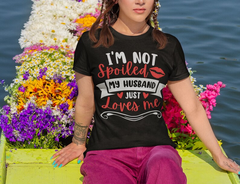 Puede incluir: Camiseta negra con el texto "I'M NOT SPOILED MY HUSBAND JUST Loves me" en rojo y blanco. El dise&ntilde;o incluye un beso de l&aacute;piz labial rojo y corazones. La persona lleva pantalones rosas y est&aacute; rodeada de flores coloridas.