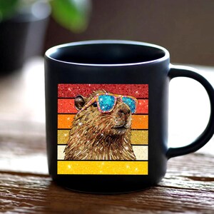 May include: A matte black ceramic mug featuring a square graphic of a brown capybara wearing bright blue glittery sunglasses. The background of the graphic consists of horizontal stripes in shades of red, orange, and yellow with a glittery finish.