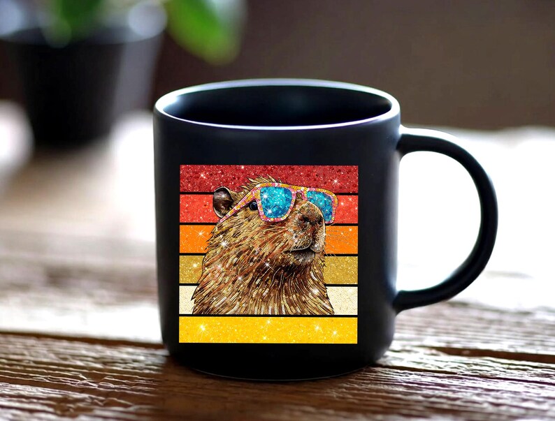 May include: A matte black ceramic mug featuring a square graphic of a brown capybara wearing bright blue glittery sunglasses. The background of the graphic consists of horizontal stripes in shades of red, orange, and yellow with a glittery finish.