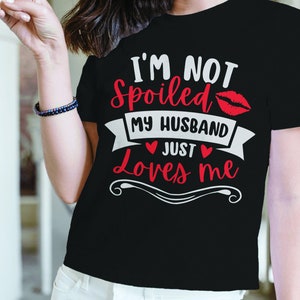 Puede incluir: Camiseta negra con el texto "I'M NOT Spoiled MY HUSBAND JUST Loves ME" en letras blancas y rojas. El dise&ntilde;o incluye un par de labios rojos y s&iacute;mbolos de coraz&oacute;n. Una prenda casual.