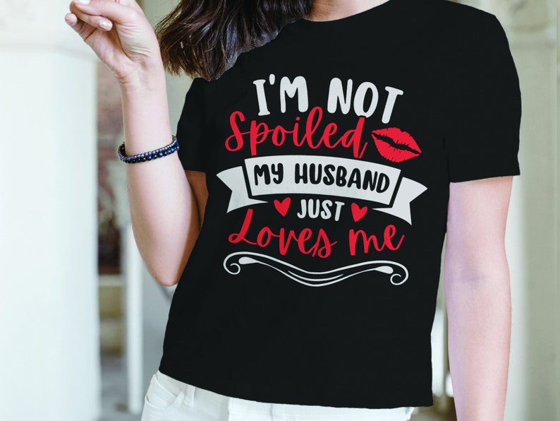 Puede incluir: Camiseta negra con el texto "I'M NOT Spoiled MY HUSBAND JUST Loves ME" en letras blancas y rojas. El dise&ntilde;o incluye un par de labios rojos y s&iacute;mbolos de coraz&oacute;n. Una prenda casual.