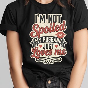 Puede incluir: Camiseta negra con el texto "I'M NOT Spoiled MY HUSBAND JUST Loves me" en letras rojas y blancas. El dise&ntilde;o incluye labios y corazones rojos, con un estilo vintage.