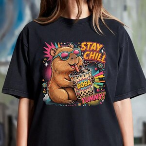 May include: A black t-shirt featuring a colorful graphic of a brown capybara wearing teal sunglasses and drinking boba tea. The design includes the text STAY CHILL, BOBA, and YUMMY, surrounded by vibrant starbursts, flowers, and cherry illustrations.