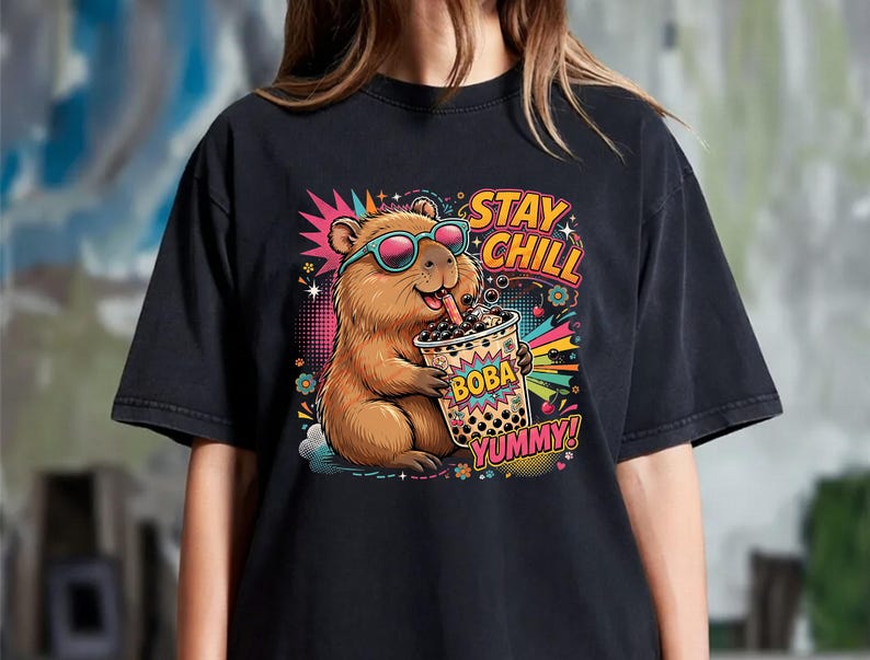 May include: A black t-shirt featuring a colorful graphic of a brown capybara wearing teal sunglasses and drinking boba tea. The design includes the text STAY CHILL, BOBA, and YUMMY, surrounded by vibrant starbursts, flowers, and cherry illustrations.