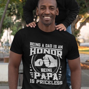 K&ouml;nnte beinhalten: Schwarzes T-Shirt mit wei&szlig;em Text, der lautet: "Ein Vater zu sein ist eine Ehre, Papa zu sein ist unbezahlbar". Der Text ist von einem Kreis umgeben, in dem sich zwei H&auml;nde einen Fauststo&szlig; geben.