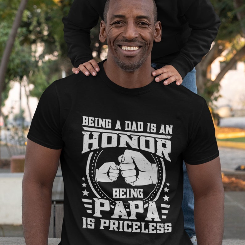 K&ouml;nnte beinhalten: Schwarzes T-Shirt mit wei&szlig;em Text, der lautet: "Ein Vater zu sein ist eine Ehre, Papa zu sein ist unbezahlbar". Der Text ist von einem Kreis umgeben, in dem sich zwei H&auml;nde einen Fauststo&szlig; geben.