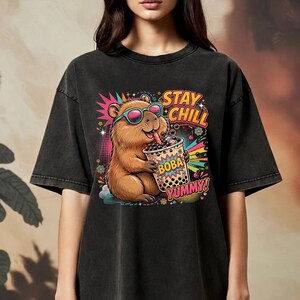 May include: A charcoal black t-shirt featuring a graphic of a brown capybara wearing pink-rimmed sunglasses and drinking boba tea. The design includes colorful starbursts, flowers, and the text STAY CHILL and BOBA YUMMY in bold, vibrant lettering.