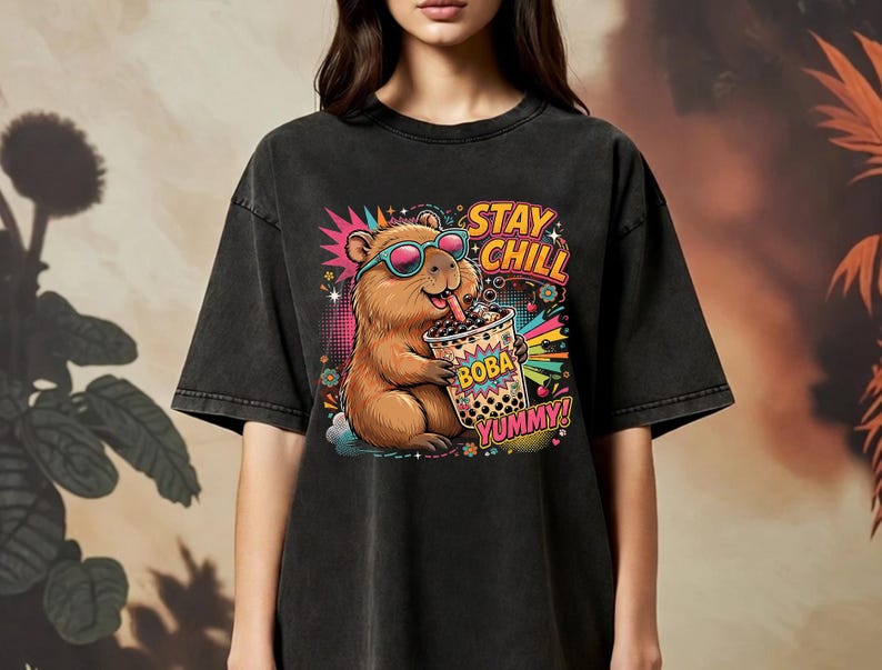 May include: A charcoal black t-shirt featuring a graphic of a brown capybara wearing pink-rimmed sunglasses and drinking boba tea. The design includes colorful starbursts, flowers, and the text STAY CHILL and BOBA YUMMY in bold, vibrant lettering.