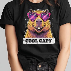 May include: A black t-shirt featuring a graphic of a capybara wearing pink heart-shaped sunglasses with a galaxy pattern inside the lenses. The capybara has brown fur and is smiling. Below the graphic, the text COOL CAPY is printed in black.