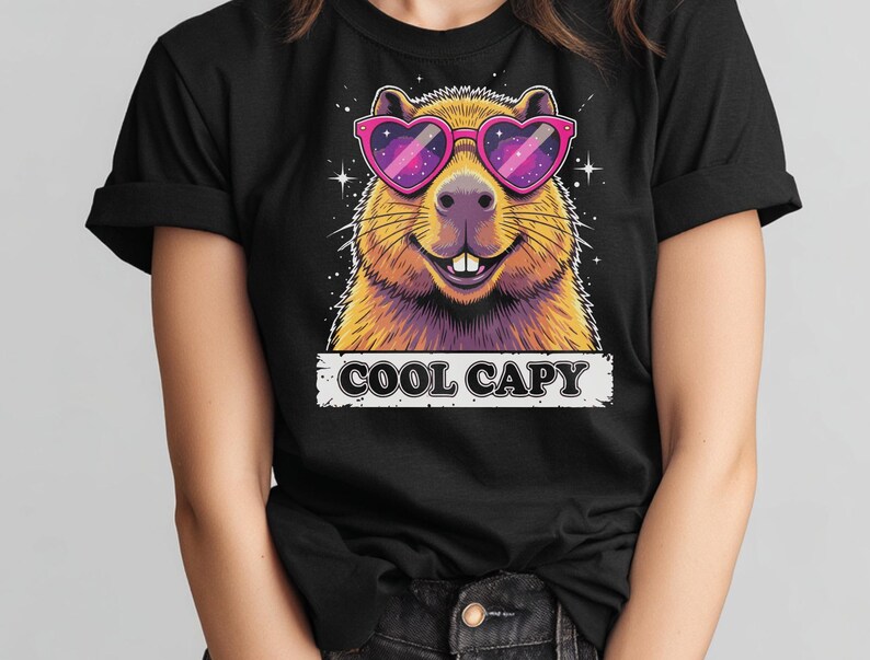 May include: A black t-shirt featuring a graphic of a capybara wearing pink heart-shaped sunglasses with a galaxy pattern inside the lenses. The capybara has brown fur and is smiling. Below the graphic, the text COOL CAPY is printed in black.