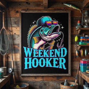 May include: A framed black poster with a cartoon fish wearing sunglasses and a hat, with the words "WEEKEND HOOKER" in blue. The fish is teal, yellow, and purple. The background is a wooden wall with fishing gear.