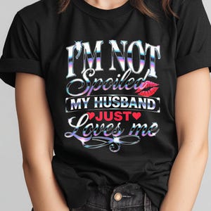 Puede incluir: Camiseta negra con el texto "I'M NOT Spoiled MY HUSBAND JUST Loves me" en una fuente hologr&aacute;fica. El dise&ntilde;o incluye un beso de labios rojos y un coraz&oacute;n rojo. Una prenda casual.