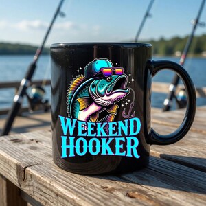 May include: Black ceramic mug featuring a colorful illustration of a fish wearing sunglasses and a cap, with the words "WEEKEND HOOKER" in turquoise. Fishing rods are visible in the background, suggesting a fishing theme.