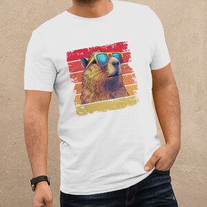 May include: A white short-sleeved t-shirt featuring a graphic print of a capybara wearing blue-tinted sunglasses. The capybara is set against a distressed, square-shaped background with horizontal stripes in shades of red, orange, and yellow.
