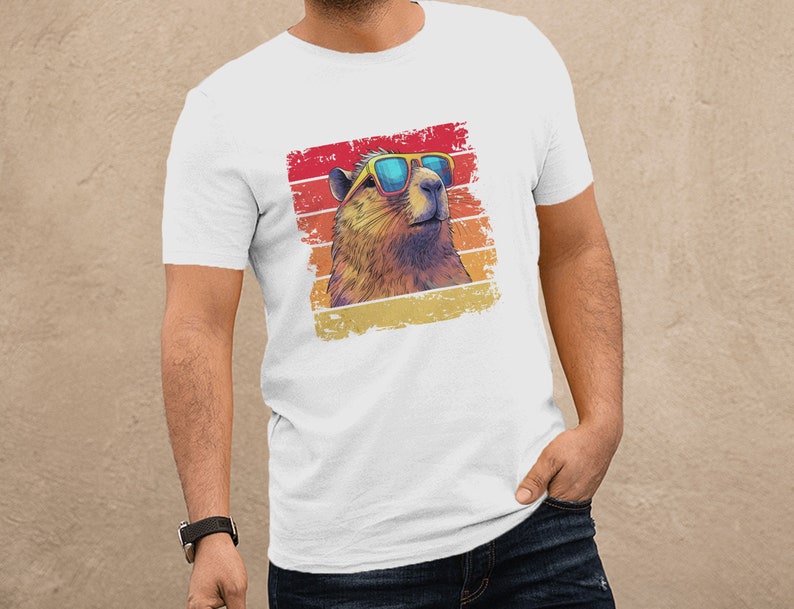 May include: A white short-sleeved t-shirt featuring a graphic print of a capybara wearing blue-tinted sunglasses. The capybara is set against a distressed, square-shaped background with horizontal stripes in shades of red, orange, and yellow.