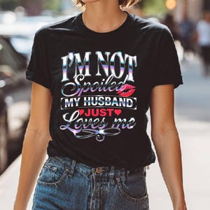 Puede incluir: Camiseta negra con el texto "I'M NOT Spoiled MY HUSBAND JUST Loves me" en una fuente hologr&aacute;fica iridiscente. El dise&ntilde;o incluye un gr&aacute;fico de labios rojos y un coraz&oacute;n. La camiseta se combina con vaqueros azules.