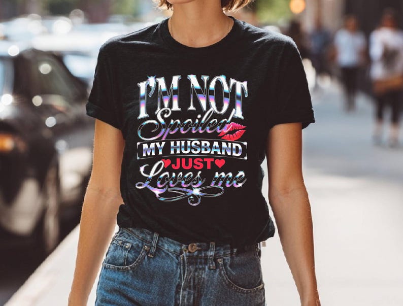 Puede incluir: Camiseta negra con el texto "I'M NOT Spoiled MY HUSBAND JUST Loves me" en una fuente hologr&aacute;fica iridiscente. El dise&ntilde;o incluye un gr&aacute;fico de labios rojos y un coraz&oacute;n. La camiseta se combina con vaqueros azules.
