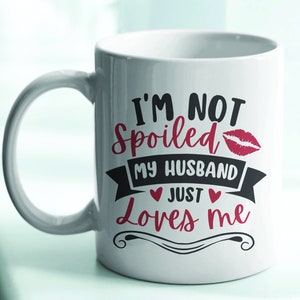 Puede incluir: Taza de cer&aacute;mica blanca con asa curva. La taza presenta el texto "I'M NOT Spoiled MY HUSBAND Just Loves me" en letras negras y rojas, con una huella de labios roja y acentos de coraz&oacute;n. La taza est&aacute; dise&ntilde;ada para parejas.