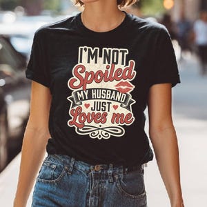 Puede incluir: Camiseta negra con el texto "I'M NOT Spoiled MY HUSBAND JUST Loves me" en un dise&ntilde;o rojo y blanco. El dise&ntilde;o incluye un par de labios rojos y un coraz&oacute;n. La camiseta se combina con unos vaqueros azules.
