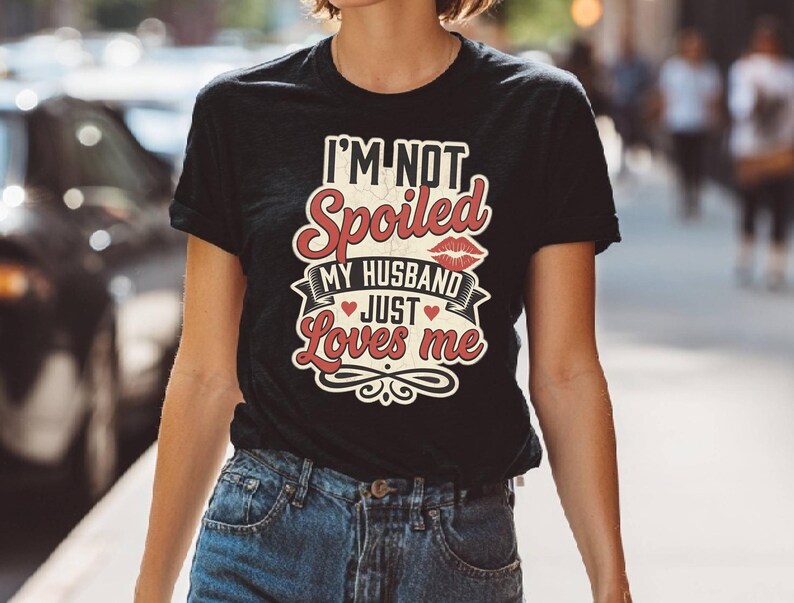 Puede incluir: Camiseta negra con el texto "I'M NOT Spoiled MY HUSBAND JUST Loves me" en un dise&ntilde;o rojo y blanco. El dise&ntilde;o incluye un par de labios rojos y un coraz&oacute;n. La camiseta se combina con unos vaqueros azules.