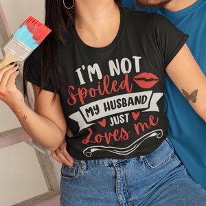 Puede incluir: Camiseta negra con el texto "I'M NOT Spoiled MY HUSBAND JUST Loves me" en blanco y rojo. El dise&ntilde;o incluye una impresi&oacute;n de labios rojos y corazones. La modelo lleva vaqueros azules.