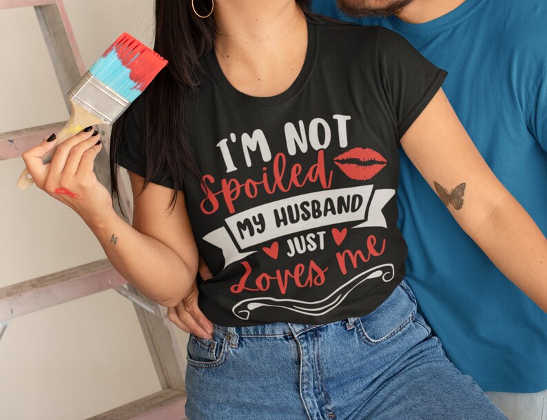 Puede incluir: Camiseta negra con el texto "I'M NOT Spoiled MY HUSBAND JUST Loves me" en blanco y rojo. El dise&ntilde;o incluye una impresi&oacute;n de labios rojos y corazones. La modelo lleva vaqueros azules.