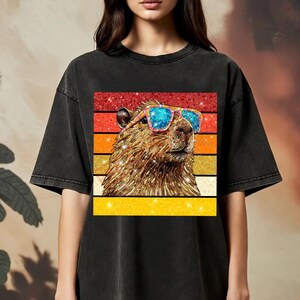 May include: A black short-sleeve t-shirt featuring a graphic print of a capybara wearing blue and pink glittery sunglasses. The capybara is set against a background of horizontal stripes in red, orange, and yellow with a sparkling glitter texture.