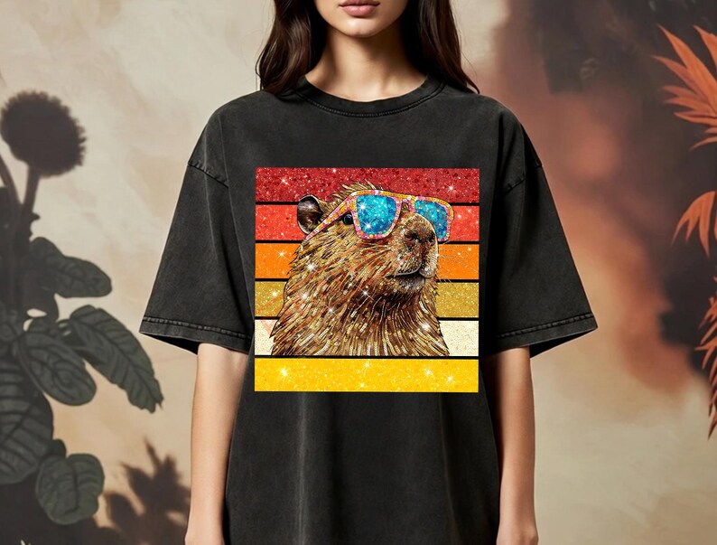 May include: A black short-sleeve t-shirt featuring a graphic print of a capybara wearing blue and pink glittery sunglasses. The capybara is set against a background of horizontal stripes in red, orange, and yellow with a sparkling glitter texture.
