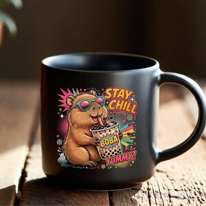 May include: A matte black ceramic mug featuring a colorful graphic of a brown capybara wearing teal sunglasses and drinking boba tea. The design includes the text "STAY CHILL" and "BOBA YUMMY" in bold, stylized lettering surrounded by vibrant, retro-inspired patterns.