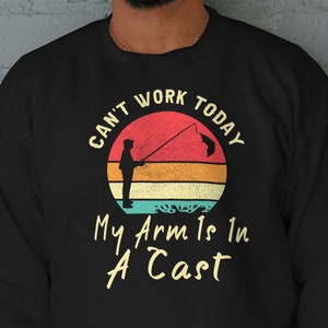 Puede incluir: Sudadera negra con un gráfico de estilo vintage de una persona pescando. El gráfico dice "Can't Work Today My Arm Is In A Cast".
