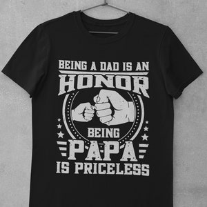 K&ouml;nnte beinhalten: Schwarzes T-Shirt mit einer wei&szlig;en Grafik, auf der steht "Ein Vater zu sein ist eine Ehre, Papa zu sein ist unbezahlbar". Die Grafik zeigt auch zwei aneinandersto&szlig;ende F&auml;uste, eine gr&ouml;&szlig;er als die andere, und Sterne.