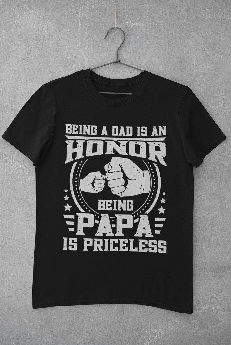 K&ouml;nnte beinhalten: Schwarzes T-Shirt mit einer wei&szlig;en Grafik, auf der steht "Ein Vater zu sein ist eine Ehre, Papa zu sein ist unbezahlbar". Die Grafik zeigt auch zwei aneinandersto&szlig;ende F&auml;uste, eine gr&ouml;&szlig;er als die andere, und Sterne.