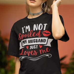 Puede incluir: Camiseta negra con el texto "I'M NOT spoiled MY HUSBAND JUST Loves me" en letras blancas y rojas. Un gr&aacute;fico de labios rojos est&aacute; encima del texto. La camiseta tiene cuello redondo y mangas cortas.