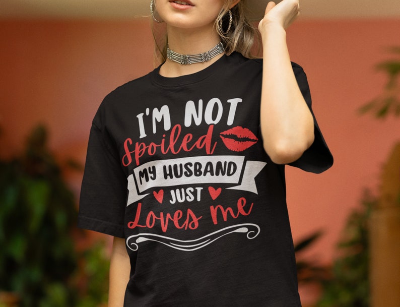 Puede incluir: Camiseta negra con el texto "I'M NOT spoiled MY HUSBAND JUST Loves me" en letras blancas y rojas. Un gr&aacute;fico de labios rojos est&aacute; encima del texto. La camiseta tiene cuello redondo y mangas cortas.