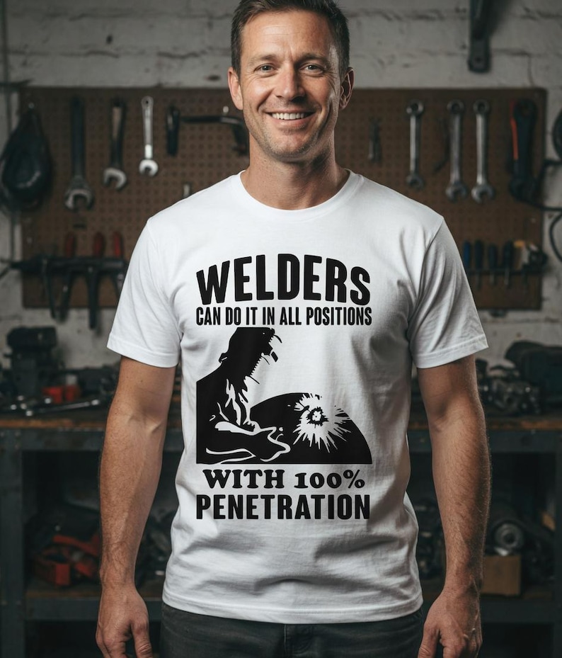 Pu&ograve; includere: T-shirt bianca con la scritta "WELDERS CAN DO IT IN ALL POSITIONS WITH 100% PENETRATION". La maglietta presenta una grafica nera di un saldatore al lavoro. La persona nell'immagine sorride.