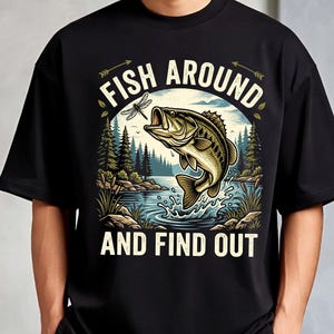 以下が含まれることがあります： 黒いTシャツで、湖から飛び出す魚と、その上にいるトンボ、周囲の松の木と葦が描かれたグラフィックイラストが特徴です。画像の上部と下部には、クリーム色のフォントで「FISH AROUND AND FIND OUT」というテキストがプリントされています。