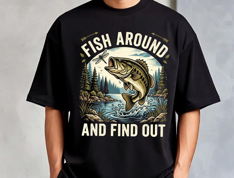 以下が含まれることがあります： 黒いTシャツで、湖から飛び出す魚と、その上にいるトンボ、周囲の松の木と葦が描かれたグラフィックイラストが特徴です。画像の上部と下部には、クリーム色のフォントで「FISH AROUND AND FIND OUT」というテキストがプリントされています。