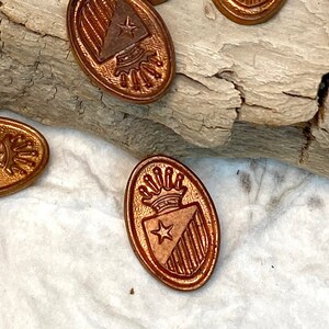 May include: A collection of nine oval-shaped, copper-toned wax seals with a design of a crown and star. The seals are scattered on a wooden surface.