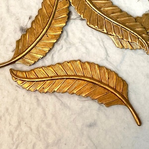 May include: A set of six gold-colored metal leaf-shaped charms. The charms are arranged in a cluster, with some overlapping. The charms are all the same size and shape, and they have a slightly textured surface.