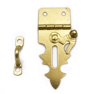 May include: A set of two brass-colored metal hardware pieces, a latch and a catch, against a white background. The latch has an ornate design with a keyhole shape and a hinged top. The catch is a small, curved piece with a loop.