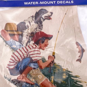 May include: A vintage water-mount decal featuring a boy fishing with a dog by his side. The decal is on a white background with a blue border. The text on the decal reads "Color Brite" and "Water-Mount Decals".