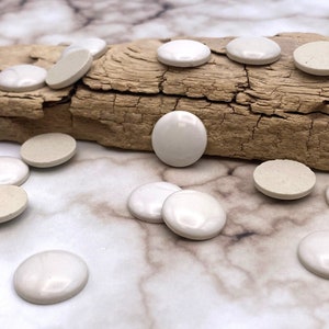 Może przedstawiać: Kolekcja okrągłych płytek ceramicznych w kolorze białym i kremowym, niektóre z połyskiem, ułożonych na drewnianym kłodzie i marmurowej powierzchni.