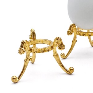 May include: Three gold-colored metal display stands with three curved legs and ornate detailing, arranged in a triangular formation. A single smooth, solid white egg-shaped object is resting securely in the center stand.
