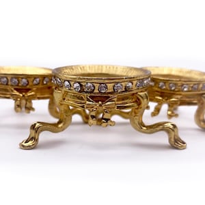 May include: Three gold-tone decorative stands with a round top and three legs. Each stand is adorned with a band of clear rhinestones and a bow-shaped design.