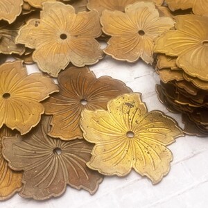 May include: A large collection of flat, brass-coloured metal flower components, each featuring five petals with embossed line details and a single centred hole. These decorative floral pieces are scattered across the surface, with some stacked.