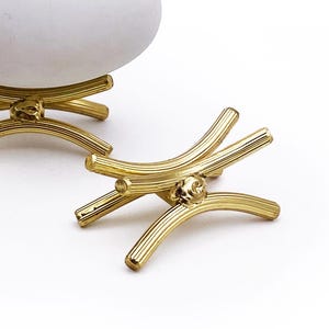 May include: Three gold-coloured metal egg holders with a unique, curved design. One holder supports a white egg, while the other two are empty. The text "*Egg NOT Included" is at the bottom.