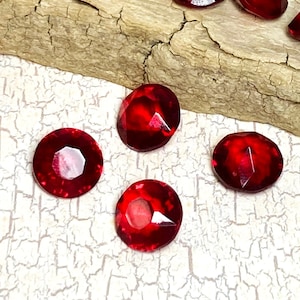 May include: A collection of faceted, deep red gemstones scattered on a piece of driftwood. The gems have a brilliant, reflective quality, catching the light. The shapes vary, including round and oval cuts. The driftwood is a light brown color.