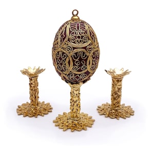 May include: A gold-colored egg stand with three legs. The stand is designed to hold a decorative egg. The egg is not included. The stand is made of metal and has a detailed design.